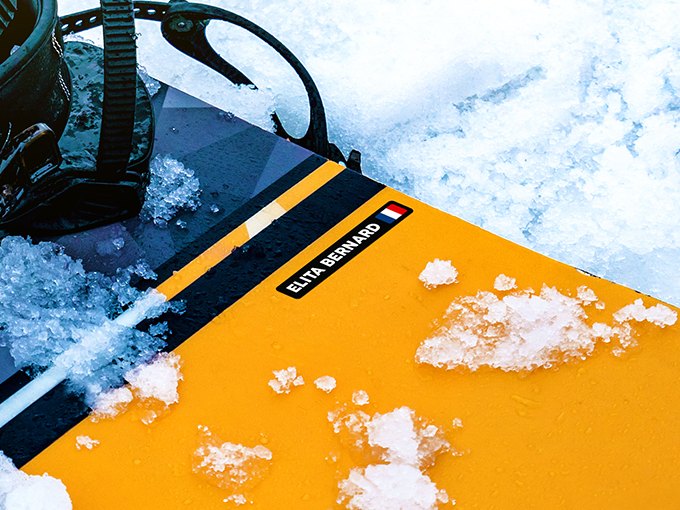 Black name sticker with French flag on a yellow snowboard in the snow